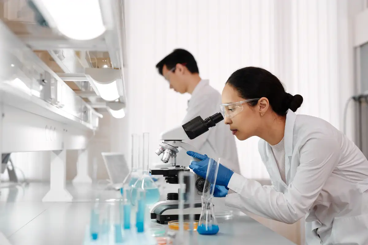 Laboratory scientist using a microscope while a colleague works nearby